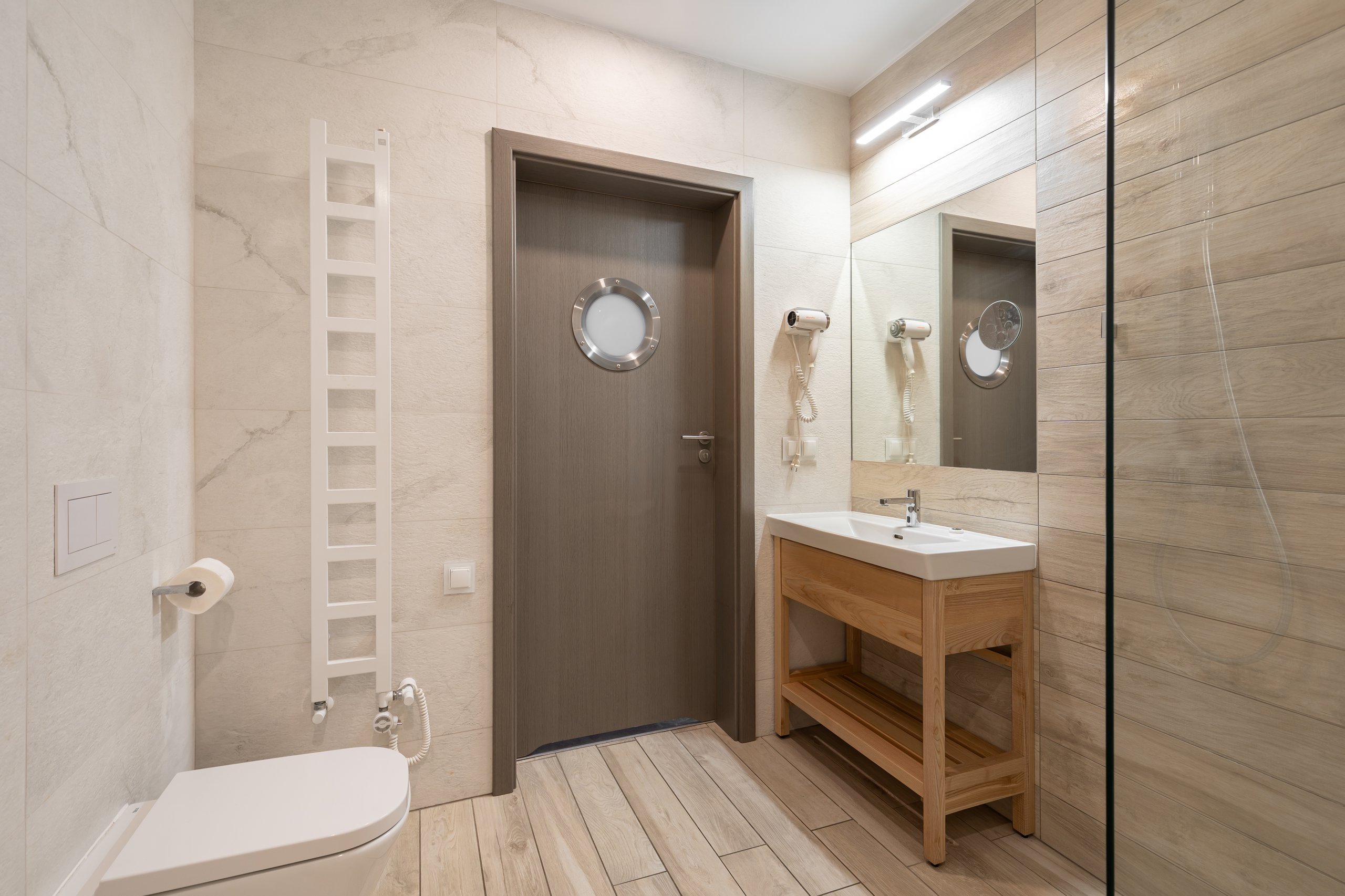 Badezimmer im Apartment in der Residenz AMBRE in Łeba – Waschbecken auf einem Holzschrank und Tür mit rundem Bullauge.
