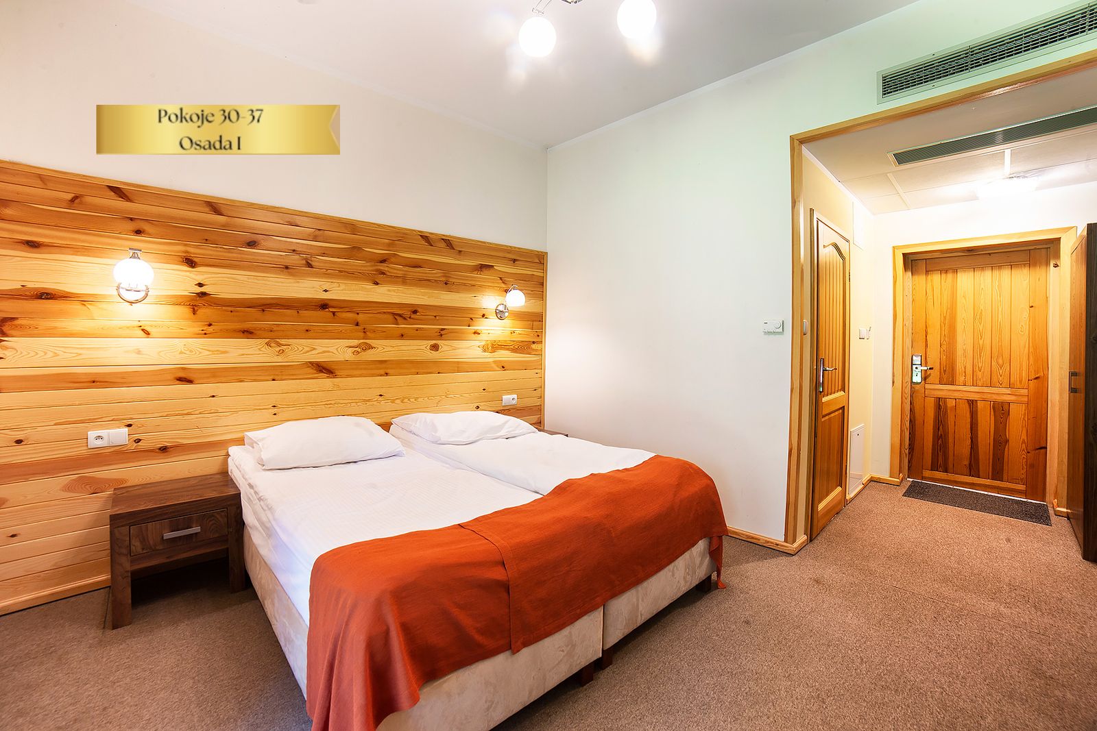 Pokój Comfort Plus w Osadzie I – łóżko z pomarańczową narzutą i drewniana ściana