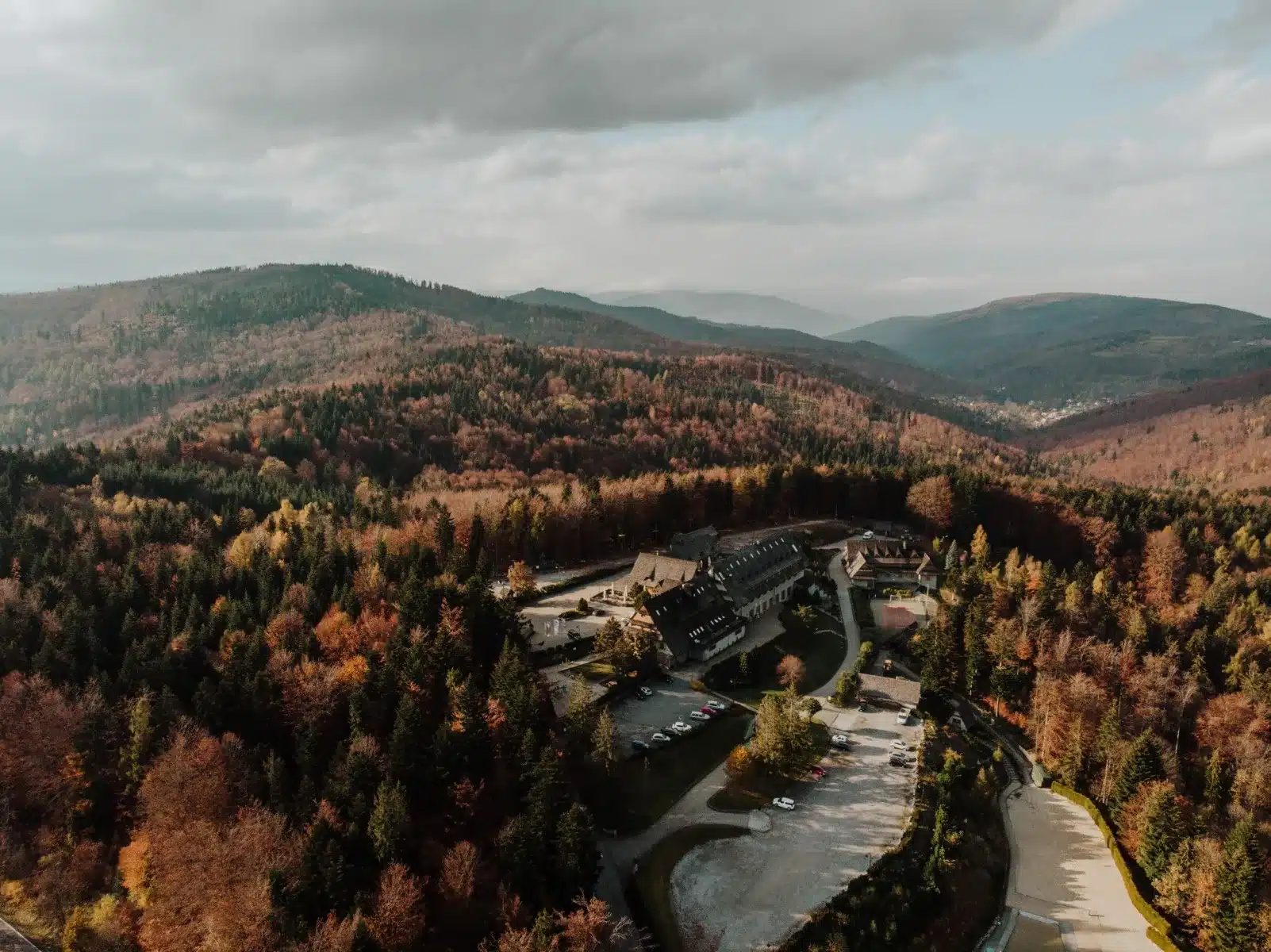 Aerial view of mountains and autumn forest with lodge complex and parking, misty overcast sky.