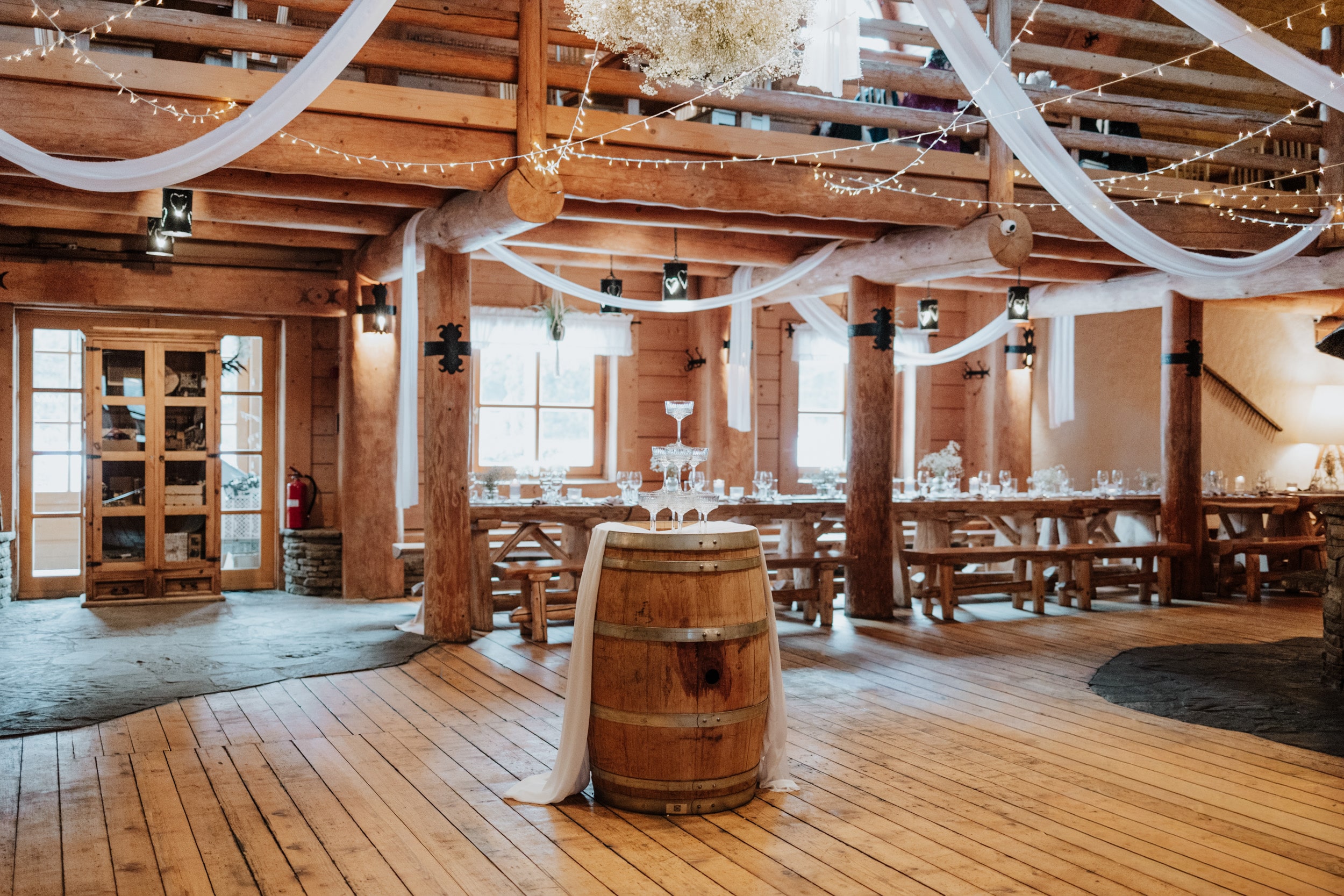 Rustic reception room with wooden barrel centerpiece, draped fabric and fairy lights, cozy atmosphere.
