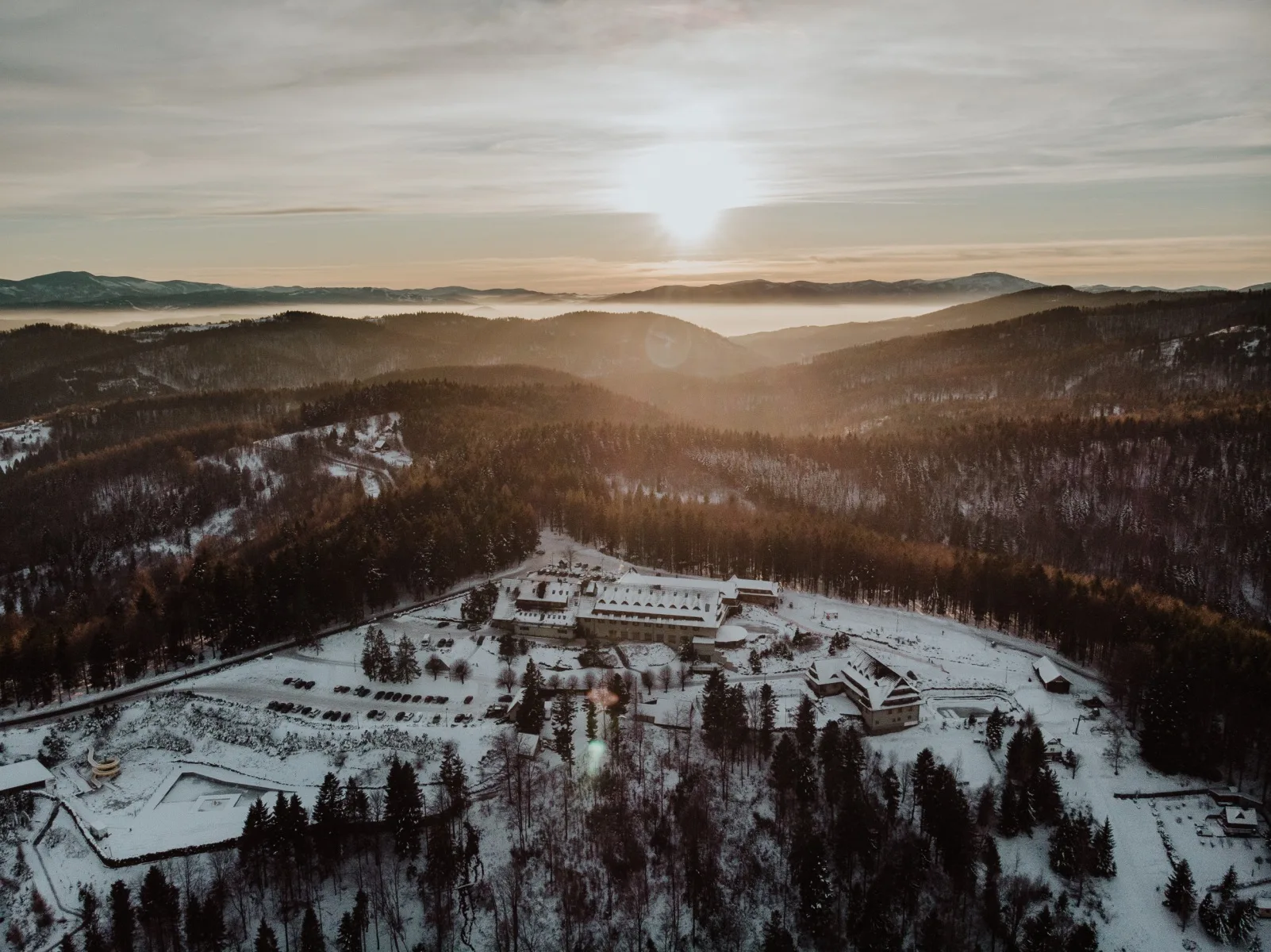 Aerial view of snow-covered resort and forest at sunrise, misty mountains and serene winter light.