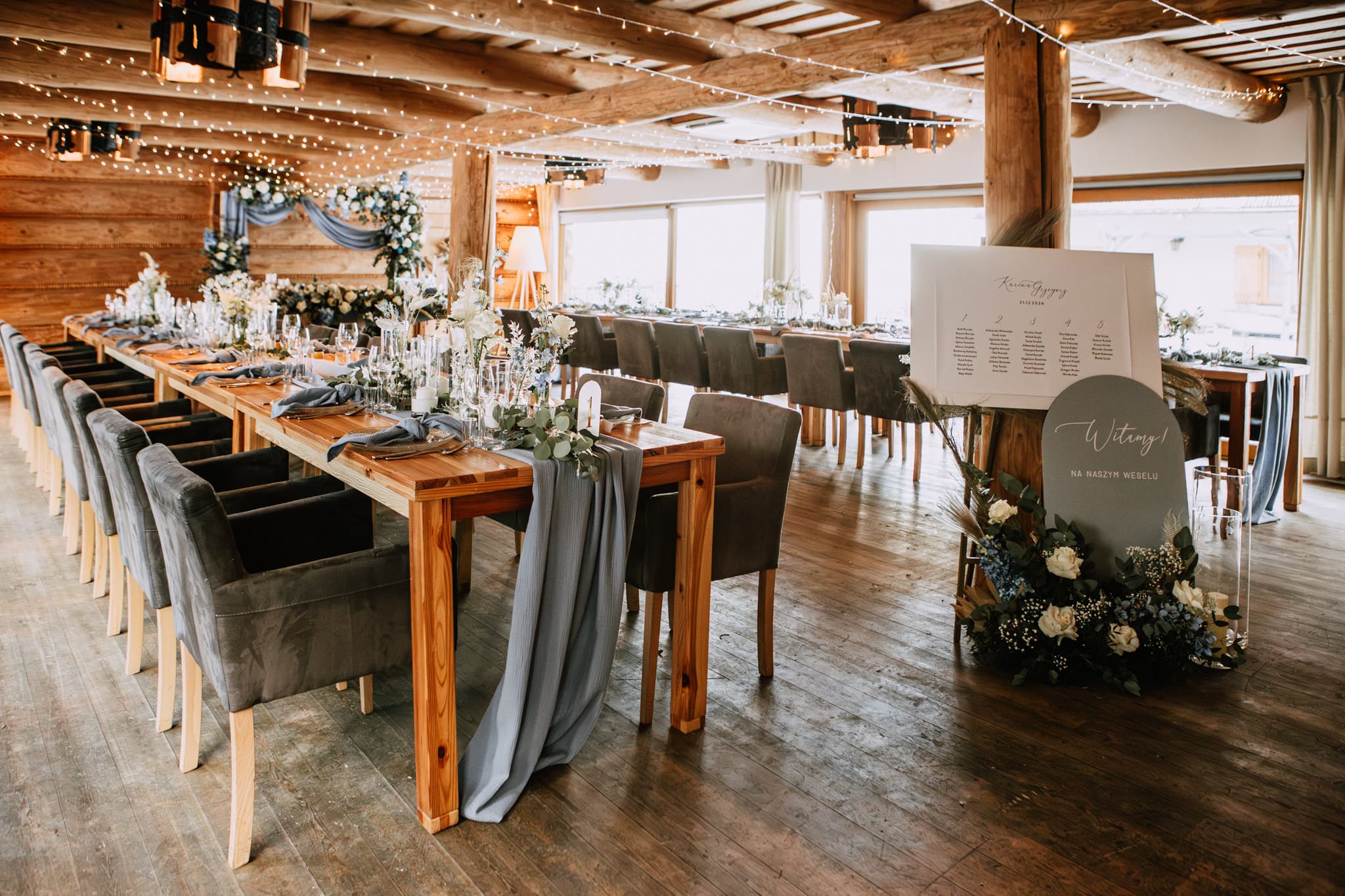 Long wooden tables set for a wedding reception in a rustic lodge, warm twinkle-light ambiance.