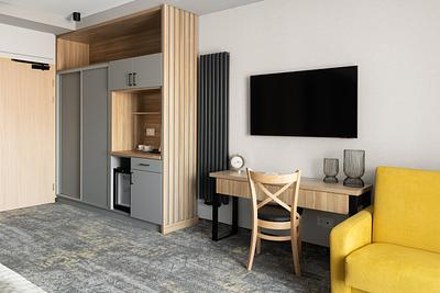 Superior-Zimmer in der Residenz AMBRE in Łeba mit Schrank, Schreibtisch und Stuhl, Fernseher und gelbem Sessel.
