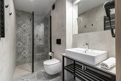 Badezimmer im Zimmer der Residenz AMBRE in Łeba – Waschbecken auf einem Metallgestell, wandhängende Toilette und Dusche mit dekorativen Fliesen.